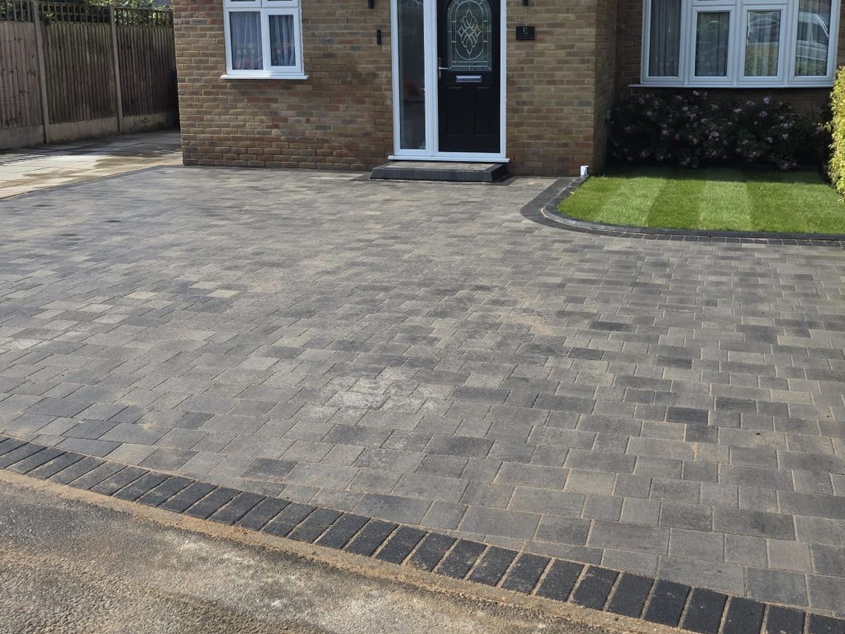 A completed grey block paved driveway in front of a house and hedge